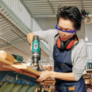 Serious young VIetnamese female carpenter in plastic goggles drilling pilot holes in wooden plank on workbench in studio